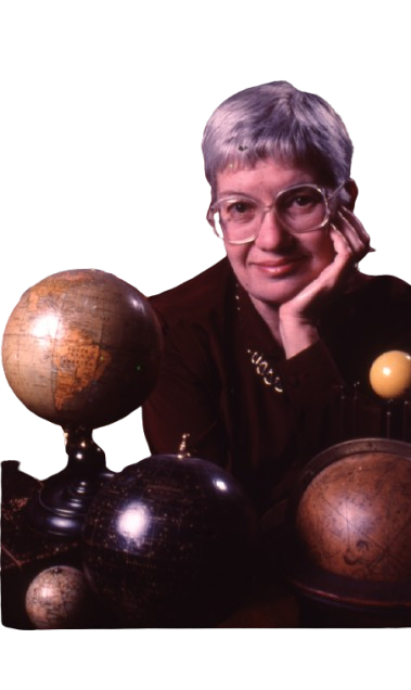 Eine Frau mit großer Brille und kurzen grauen Haaren, die hinter diversen Globen in die Kamera schaut
